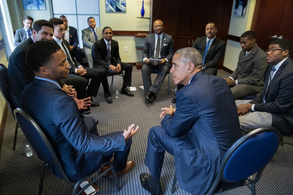 President Obama sits in a circle with students prior to the My Brother's Keeper initiative town hall hosted by ESPN at North Carolina A&T State University. All men are a range of light to deep skin tones and are dressed professionally.