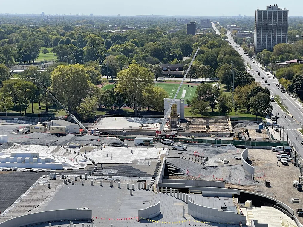 A construction site with poured concrete flooring and curved walls. Two massive cranes point in opposite directions. The site is bordered by a park with trees and fields and also a large road.