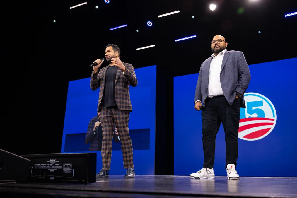 Two men are standing on a stage in an indoor auditorium. There are spotlights above them and at their feet is a black speaker. Behind the two men is a blue wall, in the middle of the wall is the Obama 15th anniversary logo. The logo has a big number 15 in the center in the color light blue. In the logo are red stripes. The first man in the picture is Kal Penn: an olive complexioned man with dark brown hair and a dark brown goatee. He is wearing a brown and black plaid suit with a black shirt underneath the suit and black leather boots. He is holding a microphone, in his right hand, to his face. The second man is Jesse Thomas: an olive complexioned man with a bald head, black framed glasses, and a black goatee. He is wearing a denim blue blazer, a white button down shirt, dark blue pants, and white sneakers. He is also holding a microphone in his right hand. 