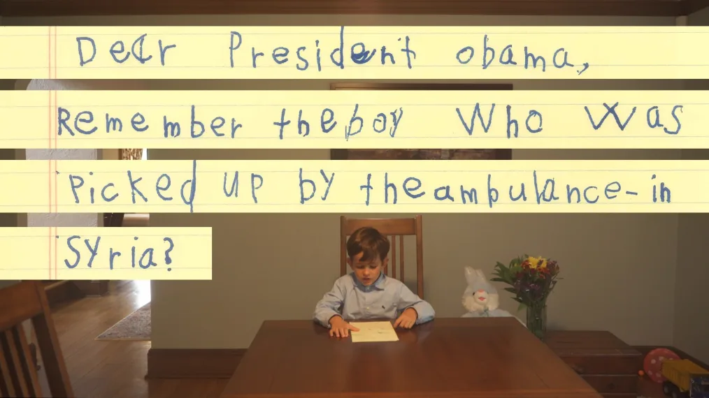 A young boy with a light skin tone sits at a table drawing. The drawing reads ¨Dear President Obama, remember the boy who was picked up by the ambulance- in syria?¨ 