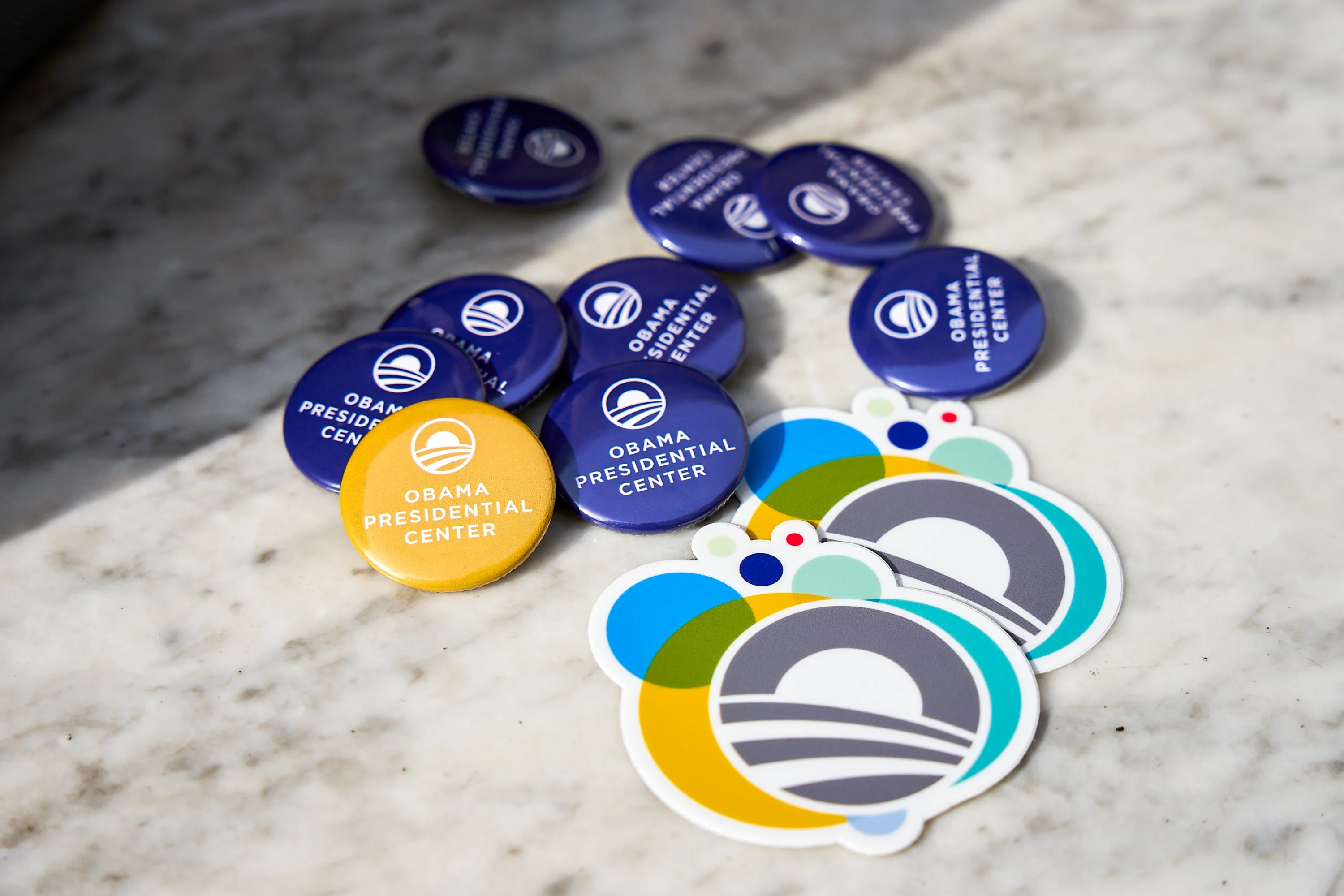 Several blue and yellow buttons that read 'OBAMA PRESIDENTIAL CENTER' and two Obama Foundation logo stickers lay across a sunny table.