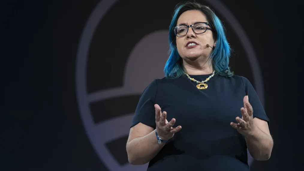 A person with shoulder-length bright blue hair wears glasses and wearable microphone moves her hands. They wear a t-shirt and gold necklace. The Obama Foundation sunrise logo can be seen behind her.
