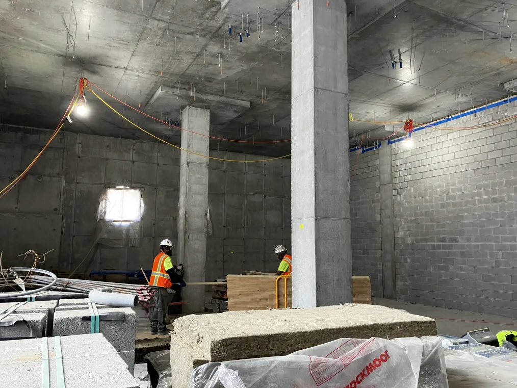 Two construction workers wearing bright orange and yellow vests appear to unload planks of wood. They stand in a cavernous room of concrete filled with insulation and other building materials.