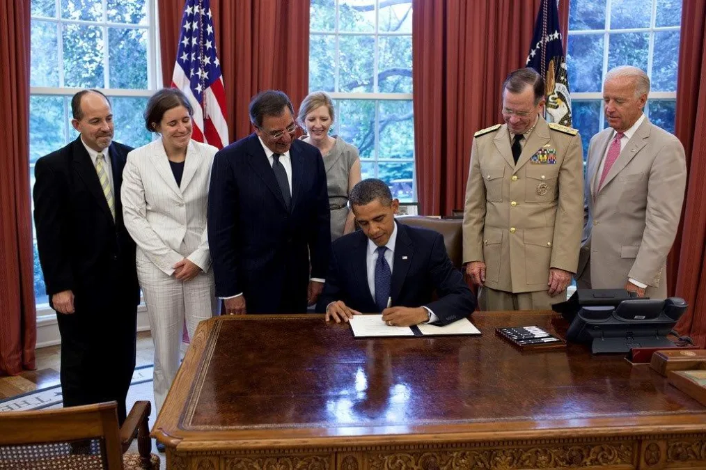 The photo is an image of President Obama certifying the repeal of Don't Ask, Don't Tell. The image is in the Oval Office. President Obama is sitting at his desk signing the certification. Standing to President Obama's far left is Brian Bond.