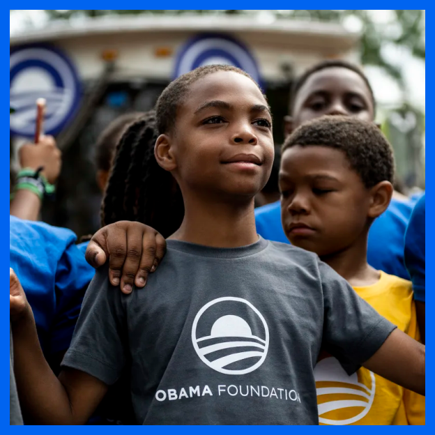 A young boy with a medium-deep skin tone stands proudly in a crowd of other young boys with medium-deep and deep skin tones. He wears a dark gray t-shirt with the Obama rising-sun logo and has his hands on the shoulders of those next to him while another puts their hand on his shoulder.