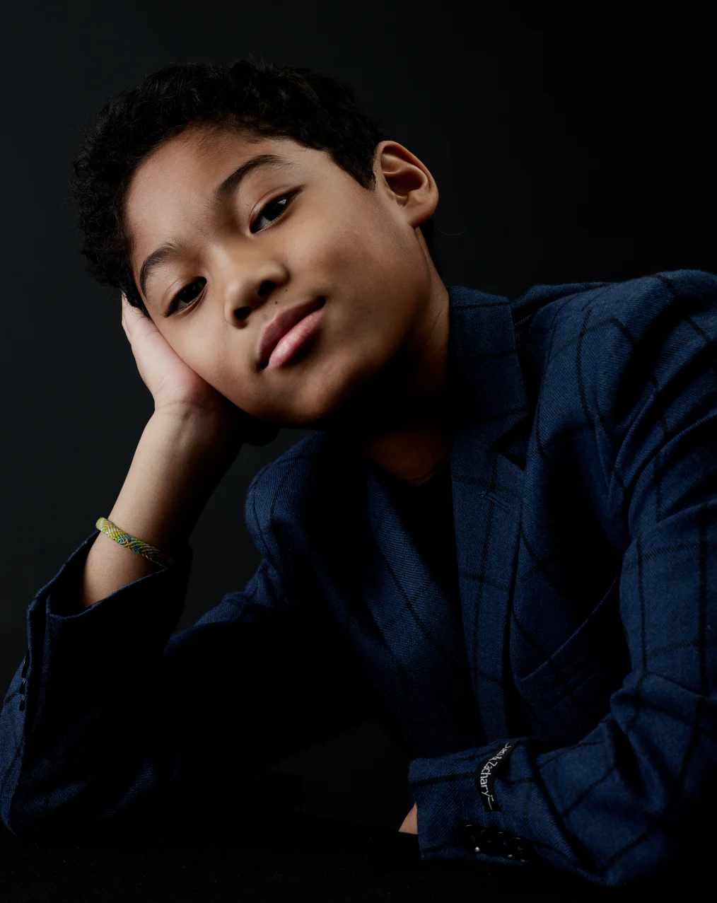 A young boy with a light skin tone rests his hand on his face as he stares into the camera. He is wearing a plaid navy suit.