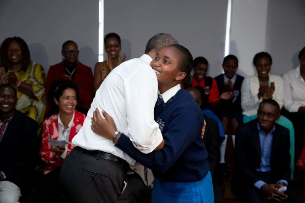 President Obama hugs a young girl with a deep skin tone and black hair infront of a crowd of diverse group of people ranging from young to old and light to deep skin tones 