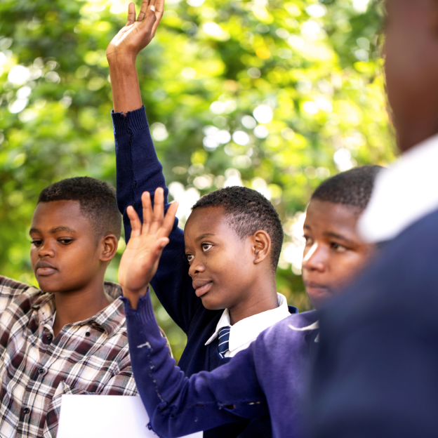 Brenda, a young Tanzanian girl with a dark skin tone, raises her hand among her classmates. The group is outside and Brenda wears a school uniform with a white collared shirt, striped tie, and sweater. They are all a range of medium to dark skin tones.