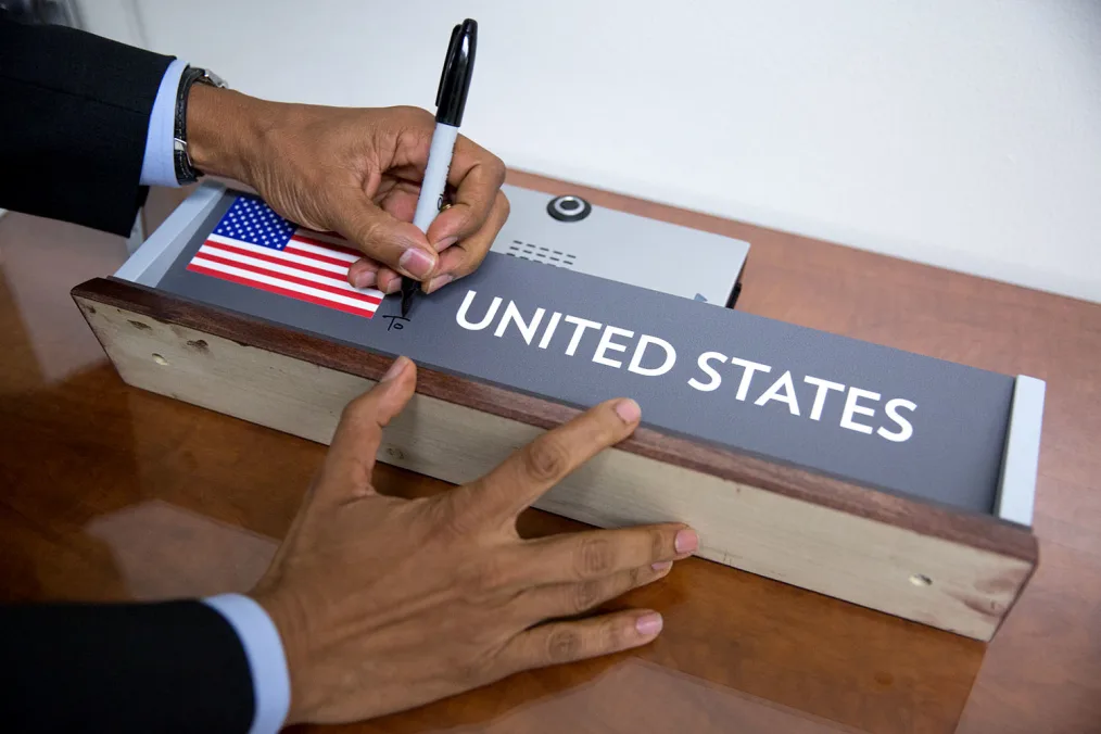 Two hands hold a wooden block with an American flag and the words "United States"on it. The hands appear to be writing the words "To" in black Sharpie marker.