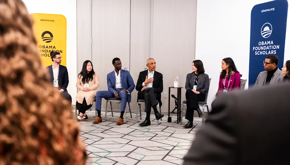 President Obama sits in a semi circle with Obama Scholars. All are dressed professionally and have a range of light to dark skin tones. Yellow and blue signs are in the background that read, “Obama Foundation Scholars.”