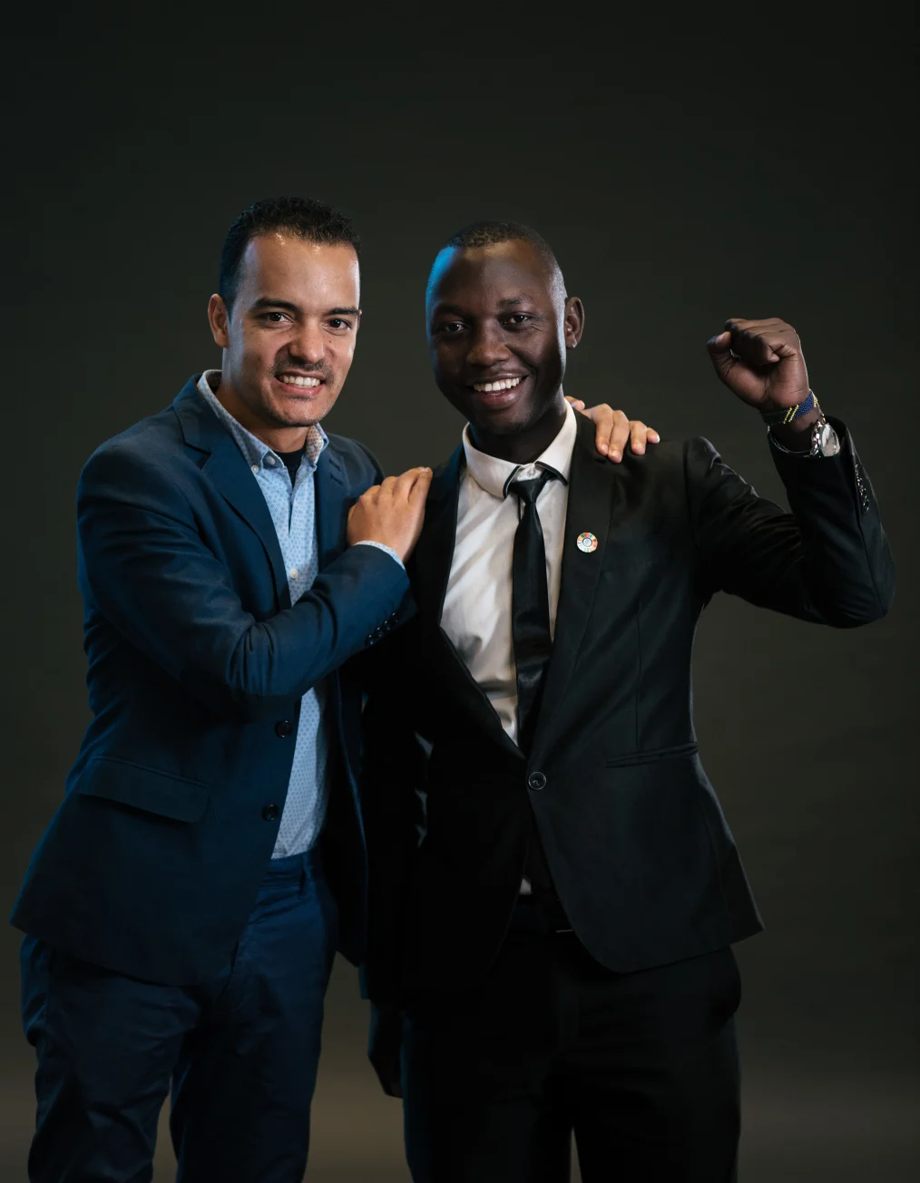 In this portrait, two men with different skin tones are shown smiling and posing
facing toward the camera with a black background.