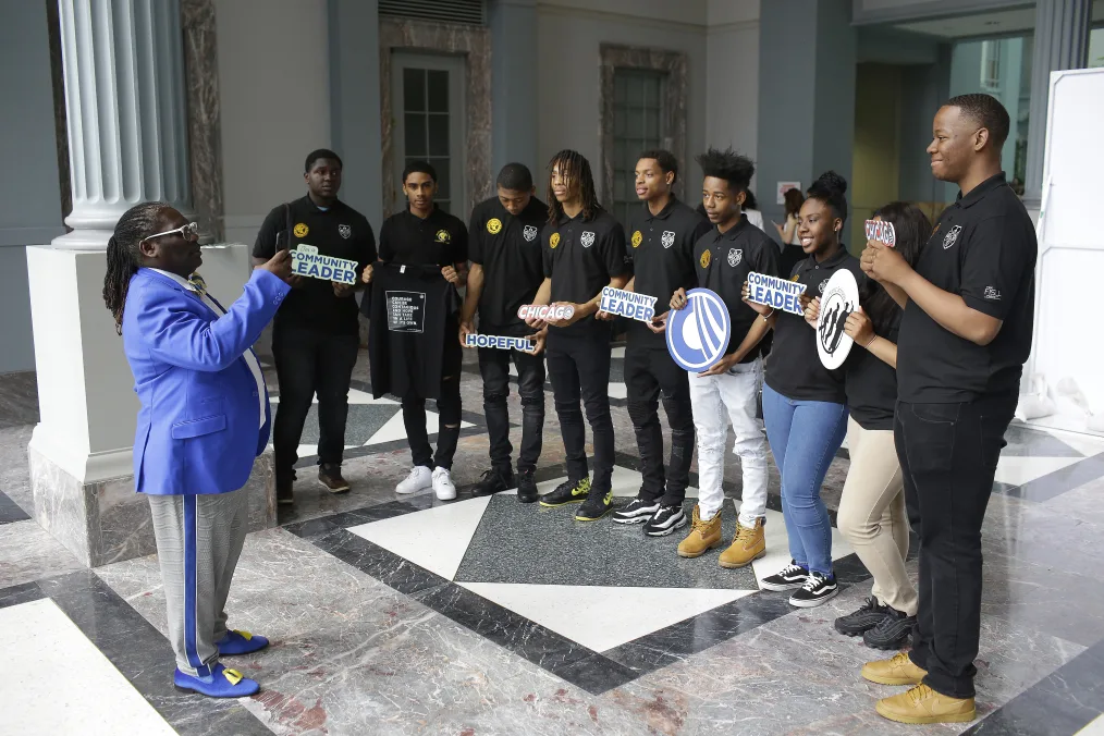 Nine people with brown skin wearing black polo shirts holding signs infront of a man with deep neutral skin wearing a blue and white suit