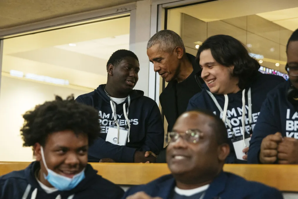 President Obama is shown shaking the hand of a young man with a deep skin tone while surrounded 
by other young men of various skin tones who are smiling.