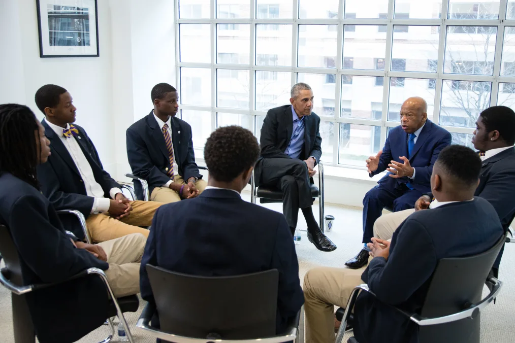 President Barack Obama and Rep.John Lewis sit in a circle with Ron Brown College Preparatory High School students. All men are a range of light to deep skin tones. 