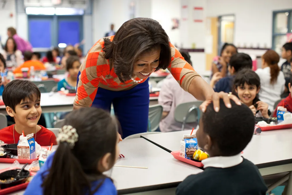 First Lady Michelle Obama, wearing a tan, red, and black patterned sweater and blue pants, reaches over a lunch table and touches the head of a young boy with a deep skin tone. Also sitting at the table are three young individuals with light-medium skin tones. There are a variety of young individuals sitting in the background blurred out.  