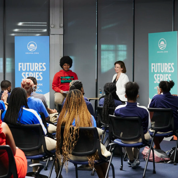 A group of students in matching blue jackest partake in a furture series meeting hosted by the Obama Foundation. The leaders of the talk are a medium-deep toned woman with an afro and glasses wearing casual attire seated next to a older light skin toned woman with medium length brown hair, glasses, and a white blazer.