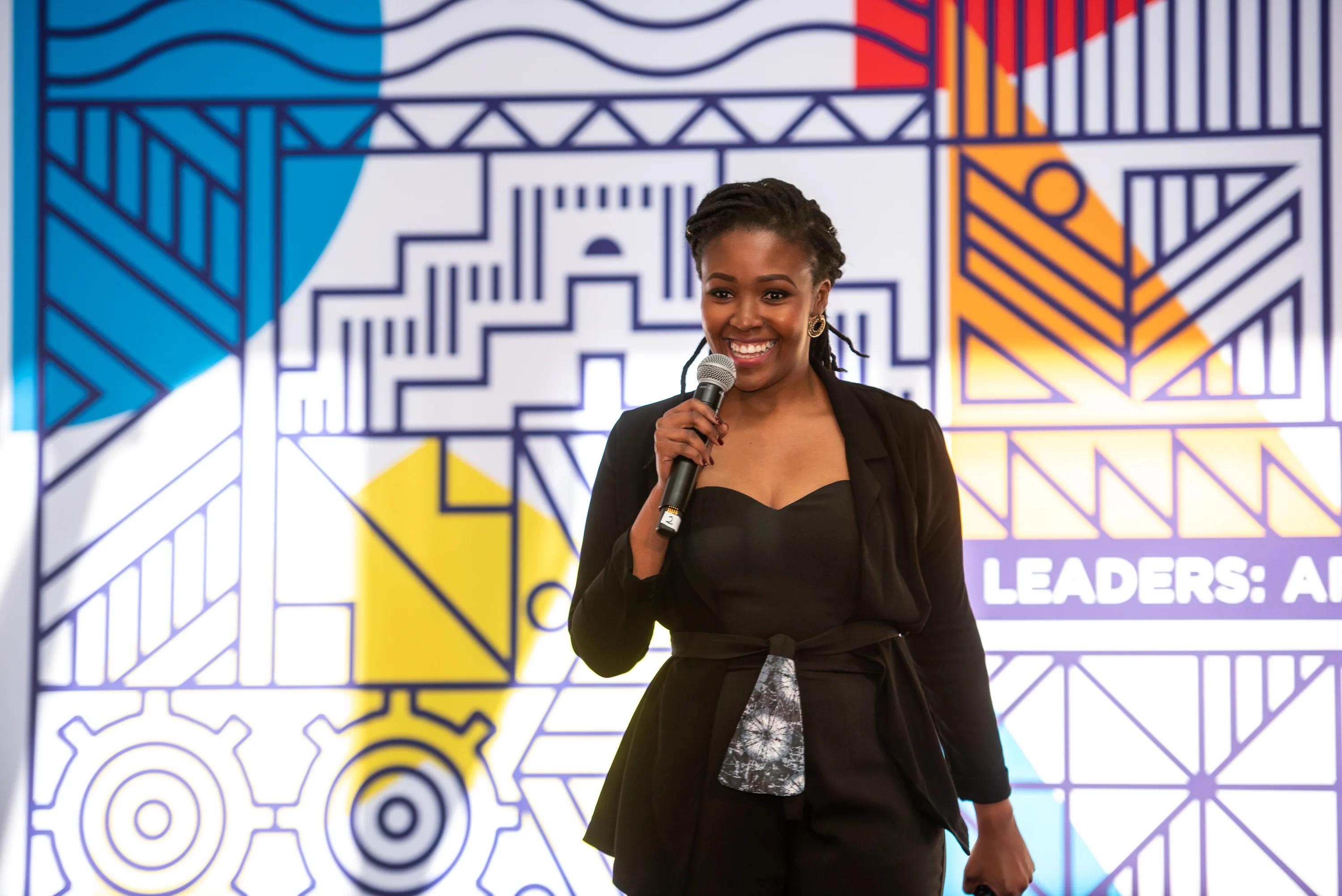 A woman with a deep skin tone holds a microphone and speaks to an unseen audience. She is smiling and has short black locs. In the background, a patterned sign reads, "Leader: Africa."