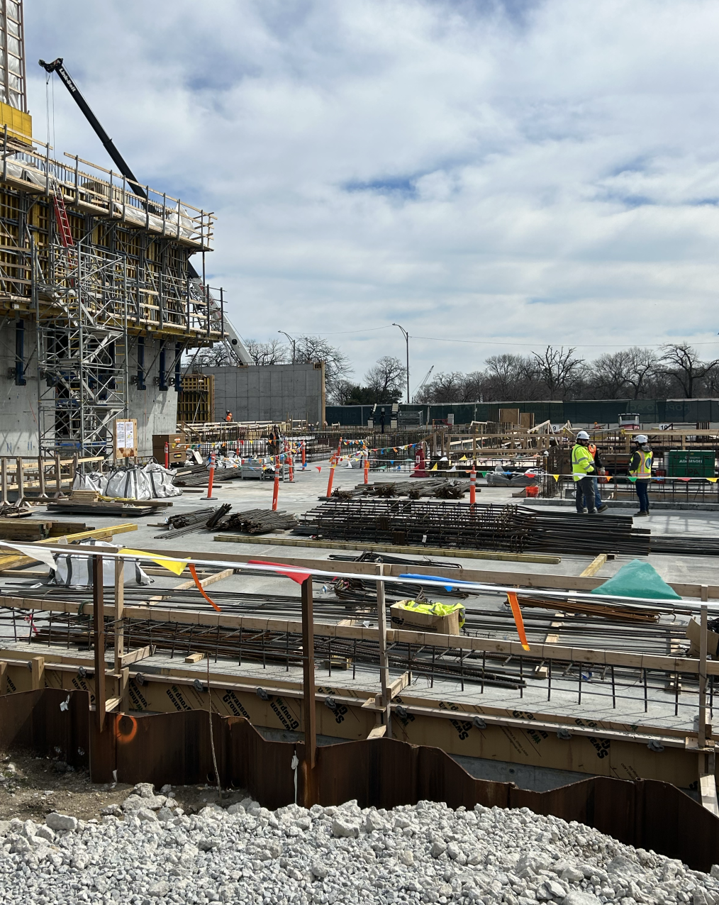 A construction site with scaffolding, concrete and rebar. Two people wear neon yellow and hard hats on the right side.