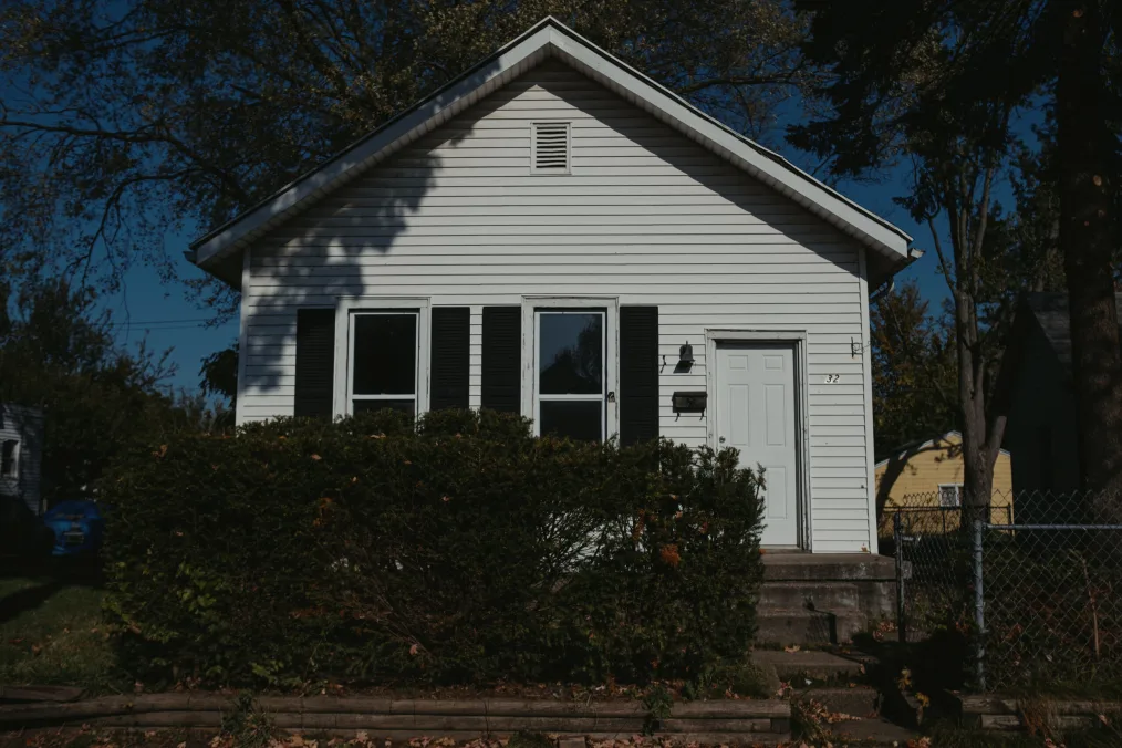 A singular, small, white, house on the side a road. In the middle slightly to the left of the picture is a farily kept square bush. On the right is a small iron gate. The house has a door on the far right and two windows on the left. There are houses on the right and left in the background. 