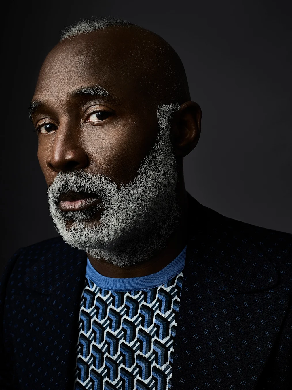 The image is a photographed portrait of Nick Cave. The background of the image is a dark gray backdrop. Nick has a deep skin tone, a short gray mohawk, and a gray goatee. He is wearing a black and blue patterned suit jacket and a blue shirt underneath.