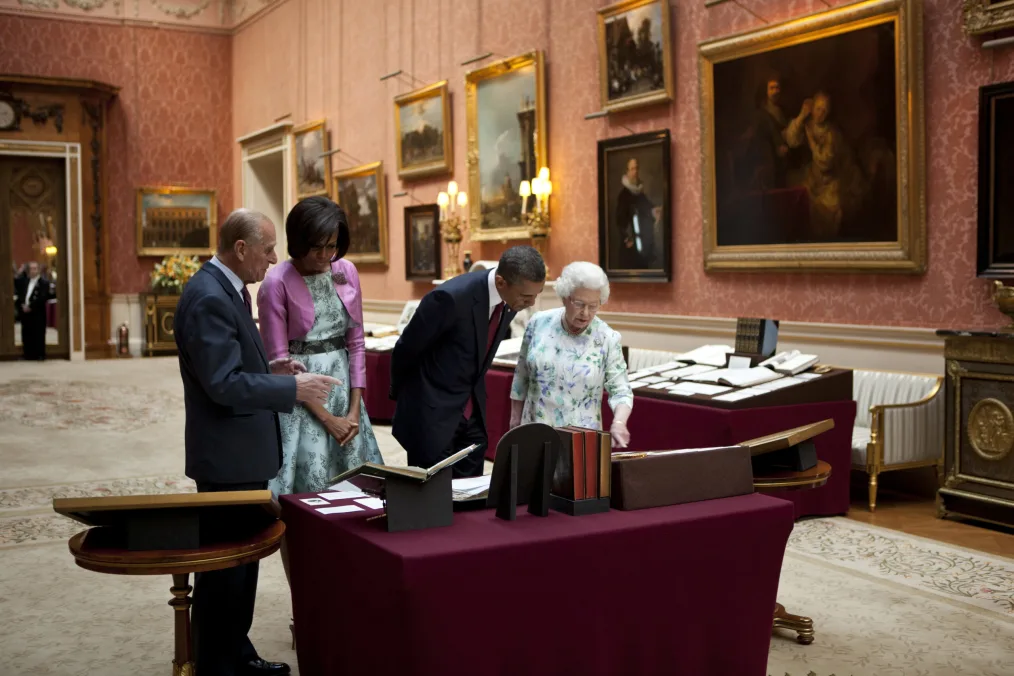 President Obama and First Lady Michelle Obama stand with a man and woman with light skin tones as they look over documents on a burgundy table. They are in a room with red patterned walls and a lot of portrait paintings. 
