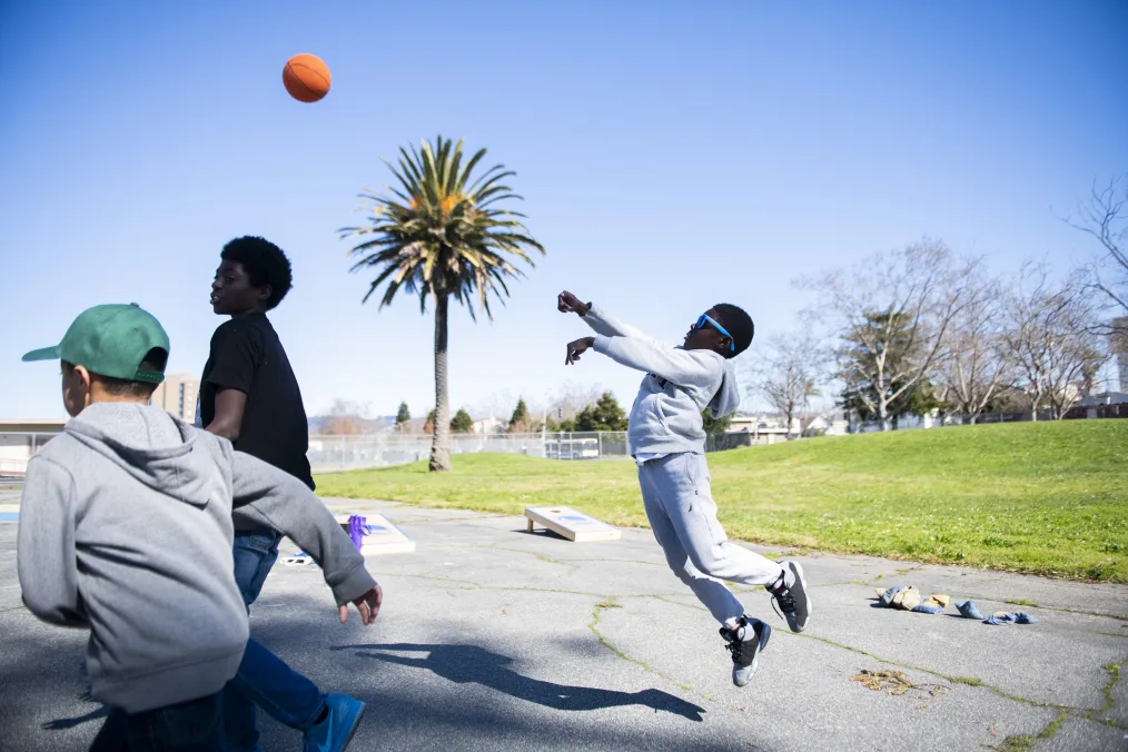 A young boy with a deep skin tone jumps in the air throwing a basketball. To the left, two more young men run. Behind them, a bright blue sky and a palm tree. 