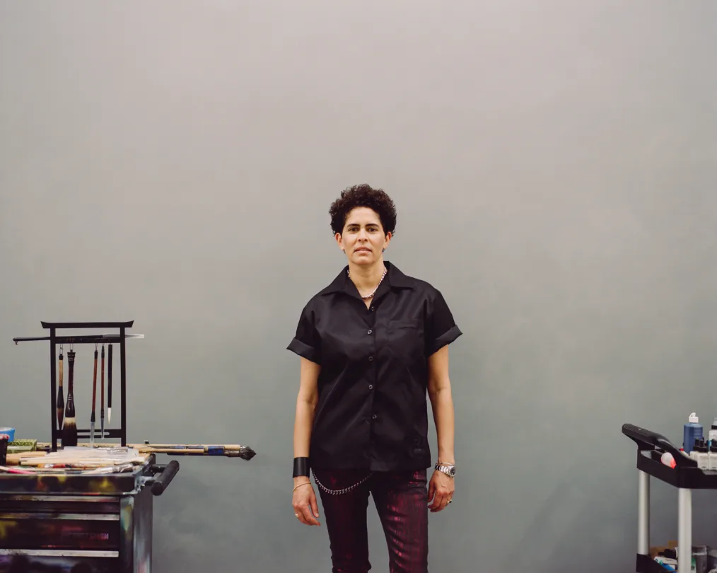 Julie Mehretu, a woman with a light skin tone and short curly black hair, stands in between two carts full of paint and art supplies. She is wearing red metallic pants. 