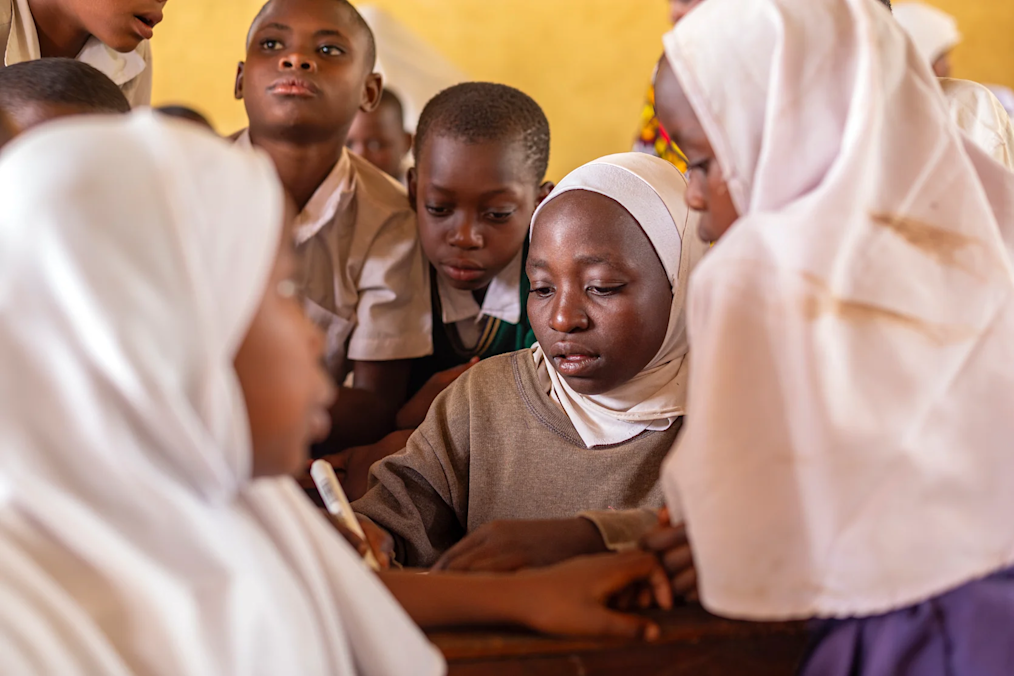 A group of girls at Secondary Education for Girls’ Advancement in Morogoro, Tanzania, with medium to dark skin tones, gather around a student writing on a pad. All are wearing uniforms and some are wearing white hijabs.