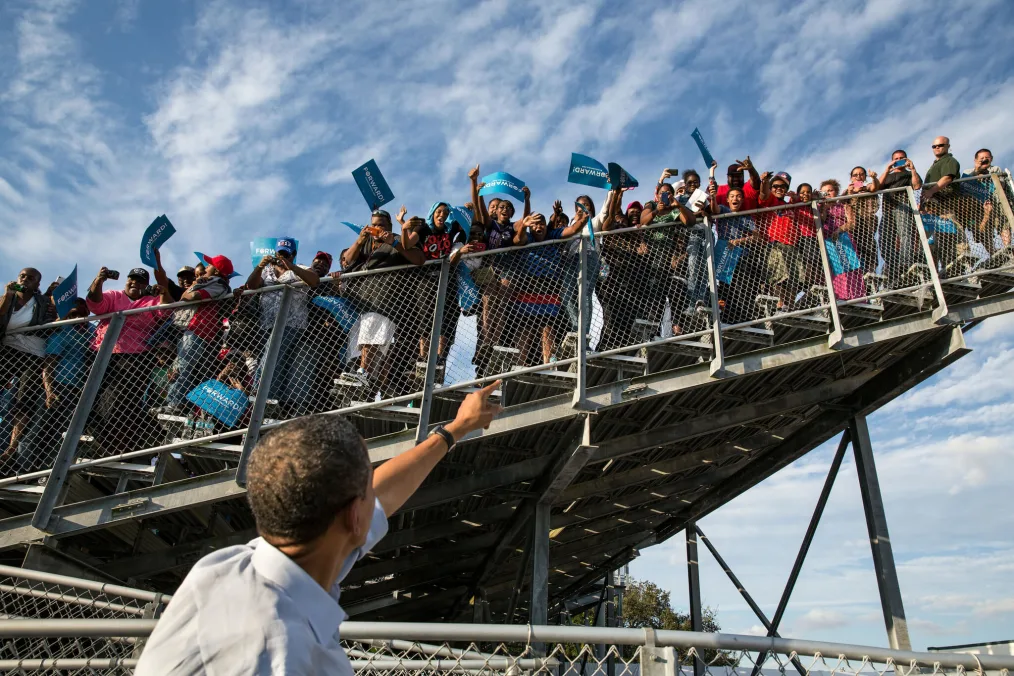 President Obama points up toward ralliers standing in bleachers.