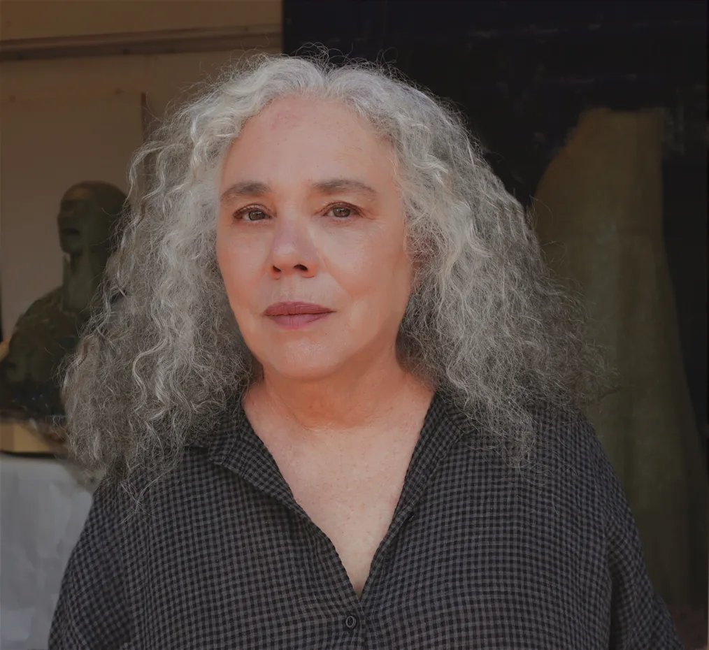 The image is a photographed portrait of Alison Saar. Alison is standing in her studio with elements of her art piece "Torch Song" in the background. Allison has a light olive complexion and long grey hair. She is wearing a checkered dark shirt with buttons down the front.