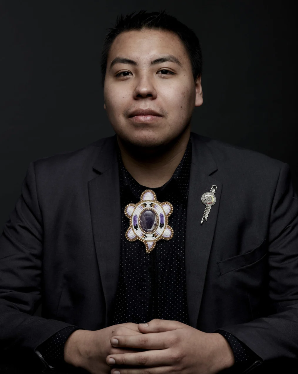 A Native American man wearing a black suit looks directly at the camera, his hands clasped in front of him. He wears an intricately beaded brooch and a lapel pin.