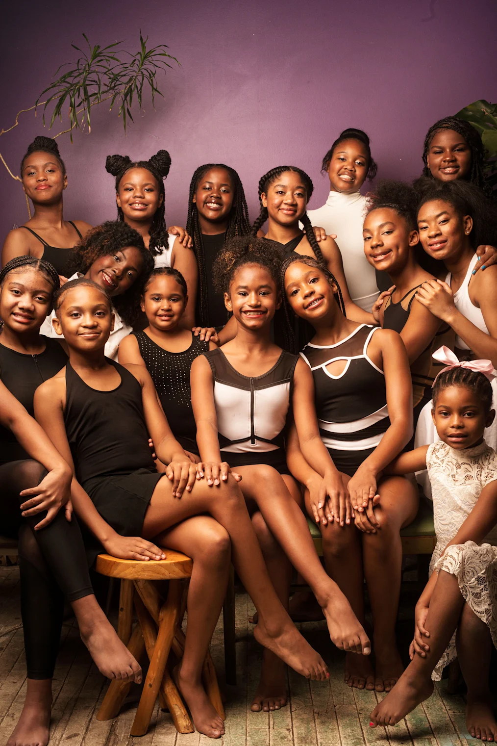 A group of young Black girls wearing dance leotards pose for a photo against a purple background. 