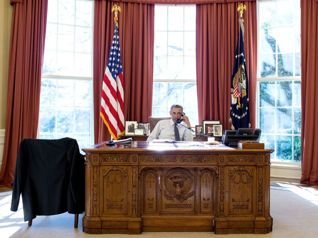 President Barack Obama sits at a large, ornate wood desk in the oval office. He is on the phone. Behind him are two flags, long red drapes and large windows.