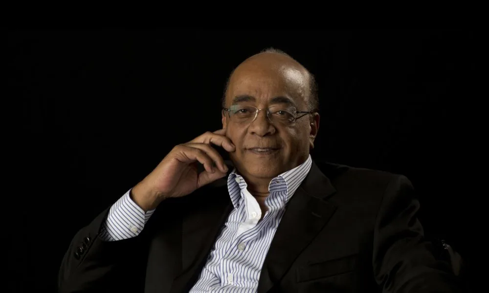 A man with a medium-deep skin tone, glasses, a blue and white striped button-up shirt, and a black blazer sits and smiles in front of a solid black background. 