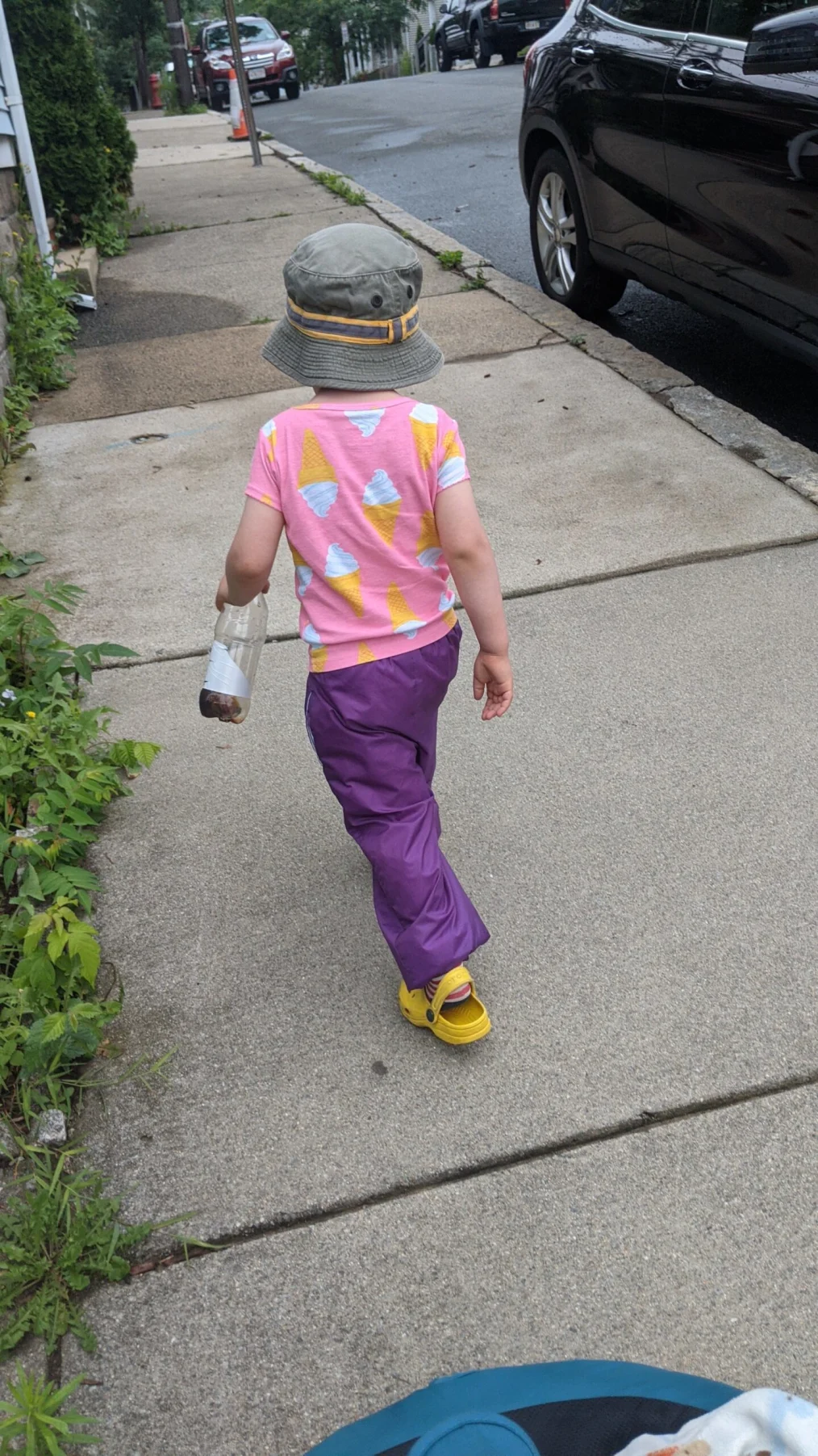 A toddler walks ahead of her mom, wearing a pink shirt on and a bucket hat.