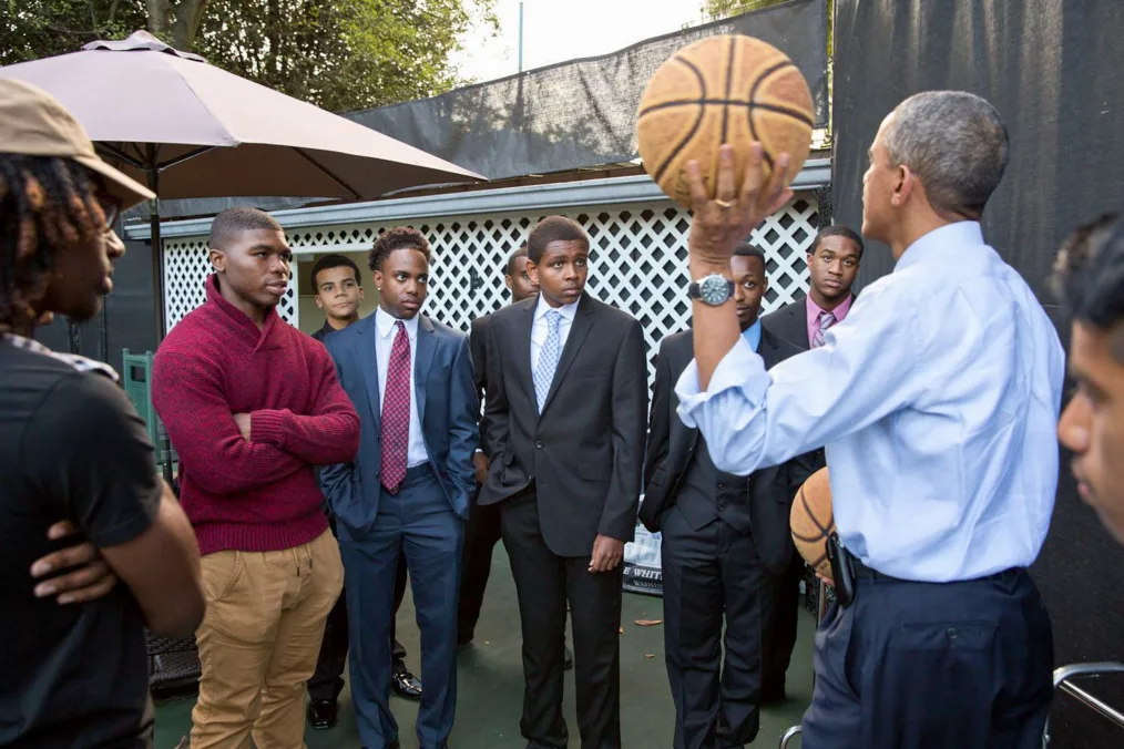 President Obama holds a basketball as he speaks to a group of young men. All men are a range of light to deep skin tones and are dressed professionally. 