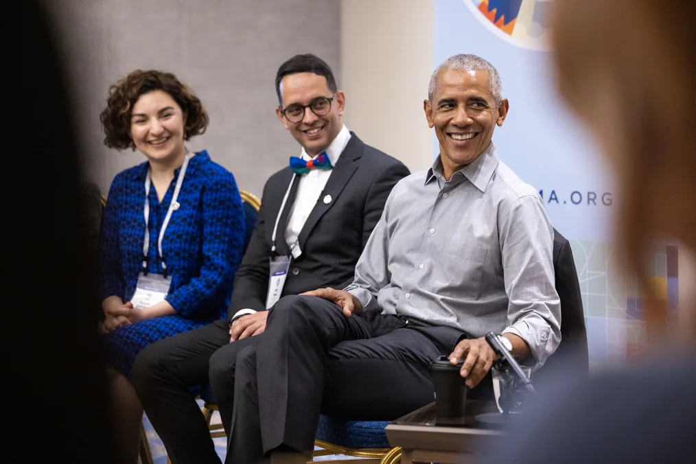 President Obama smiles as he listens to a group of people of varying skin tones talk. To his right are two members of the Obama Leaders Europe cohort of varying skin tones. They are also smiling. In the background there is a banner of the Obama Leaders Europe medallion.