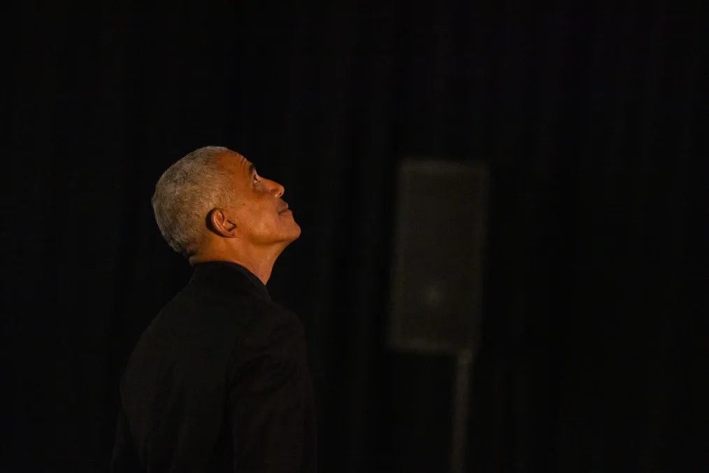 The image is a photograph of Barack Obama looking at the Power of Words prototype. The background of the image is dark and the only person in the photo is Barack Obama. He is looking up at the Power of Words prototype (not in the picture) and smiling with no teeth. He is wearing a black blazer. 