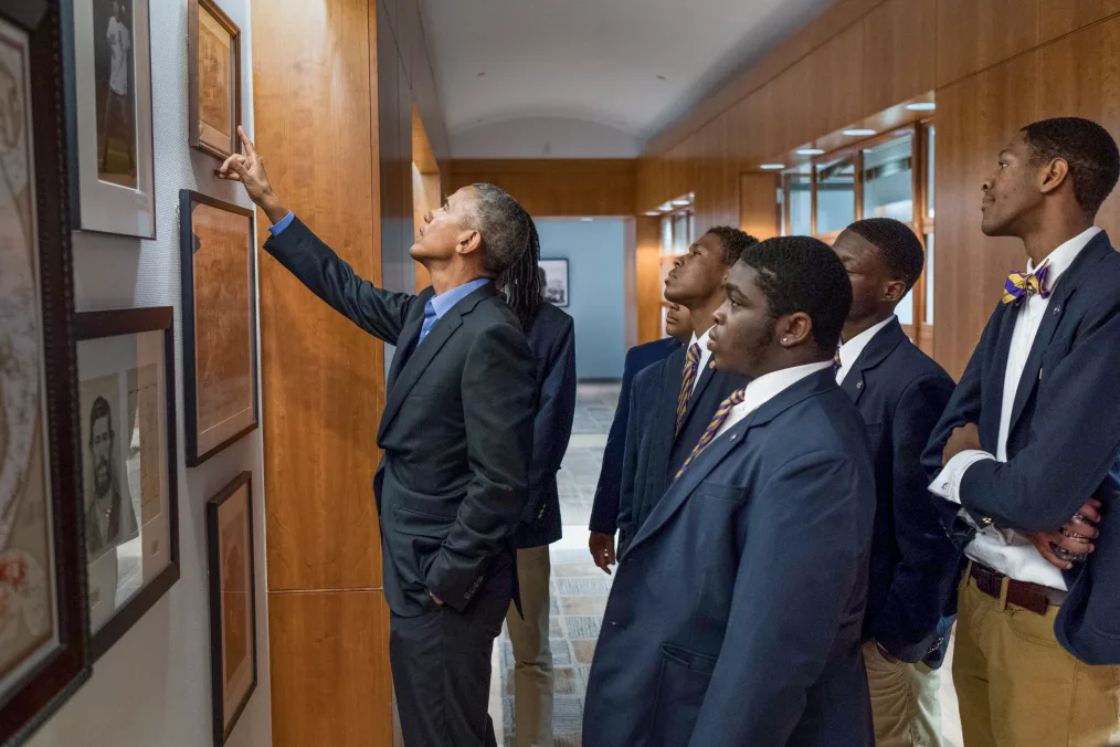 President Obama points to a photo on a wall. Behind him a group of young men with a range of medium to deep skin tones look on. 