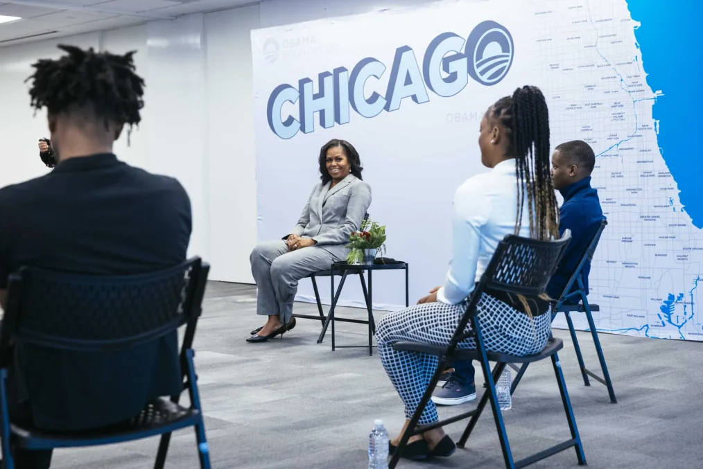 Michelle Obama smiles during a circle group meeting with young men and women. They are evenly spaced out and seated wearing formal attire. In the background, there is a graphic on a wall of the word Chicago and a mini map of the city.