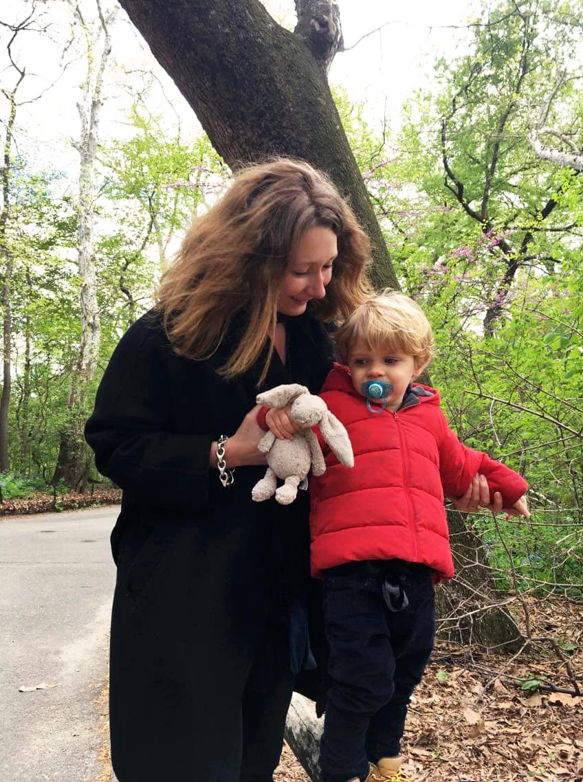 An image of a woman with a light skin tone wearing a black dress balances a young boy with a light skin tone wearing a red coat and holding a toy bunny outdoors on a wooden railing. 