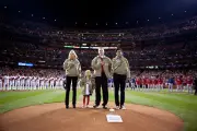 First Lady Michelle Obama wearing a tan jacket and black pants stands on the grass amongst three individuals with light skin tones wearing tan jackets and black pants. In the background there are baseball teams and a stadium full of people with a variety of skin tones. 