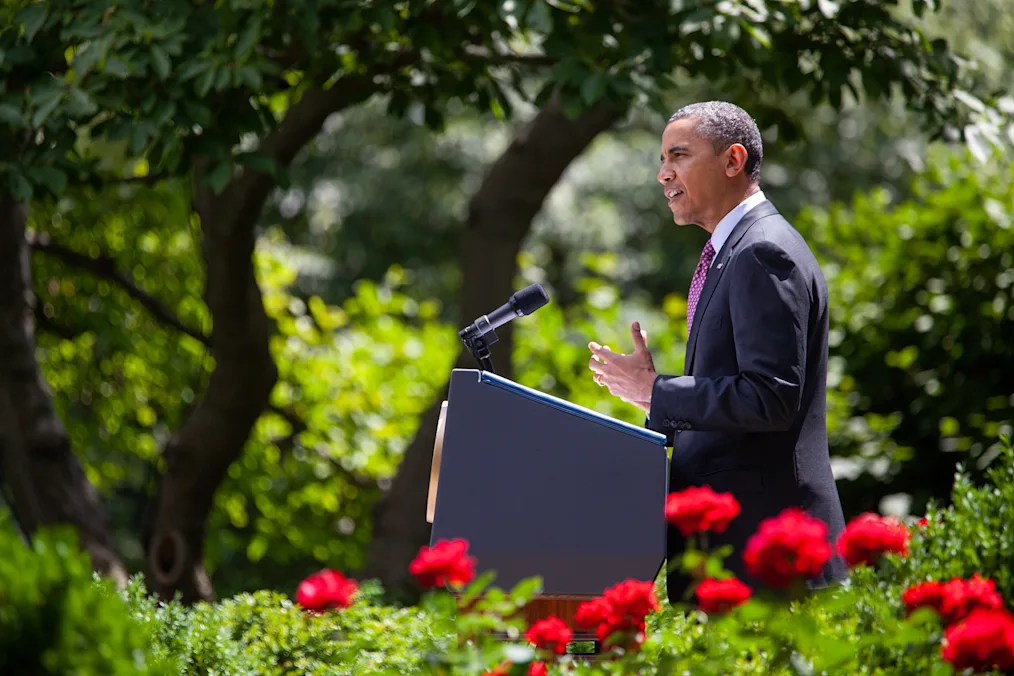 An image of President Barack Obama speaking in the Rose Garden of the White House. President Obama has a medium deep skin complexion and close cut hair that is black with small sprinkles of gray. He is wearing a dark suit with a red tie with blue polka dots. He is standing behind a black podium with a microphone facing him. Behind him are trees with green leaves. In the front of the image are several rose bushes. The roses on the bushes are red. It is sunny and daylight outside.