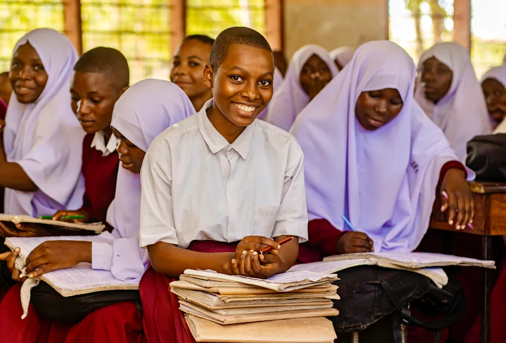 In the center of the photo is a young girl with a dark skin tone and no hair. She is smiling as she rests her hands on top of several books. She is surrounded by a group of girls with dark skin tones reading books. Most of the girls in the background are wearing purple hijabs. 