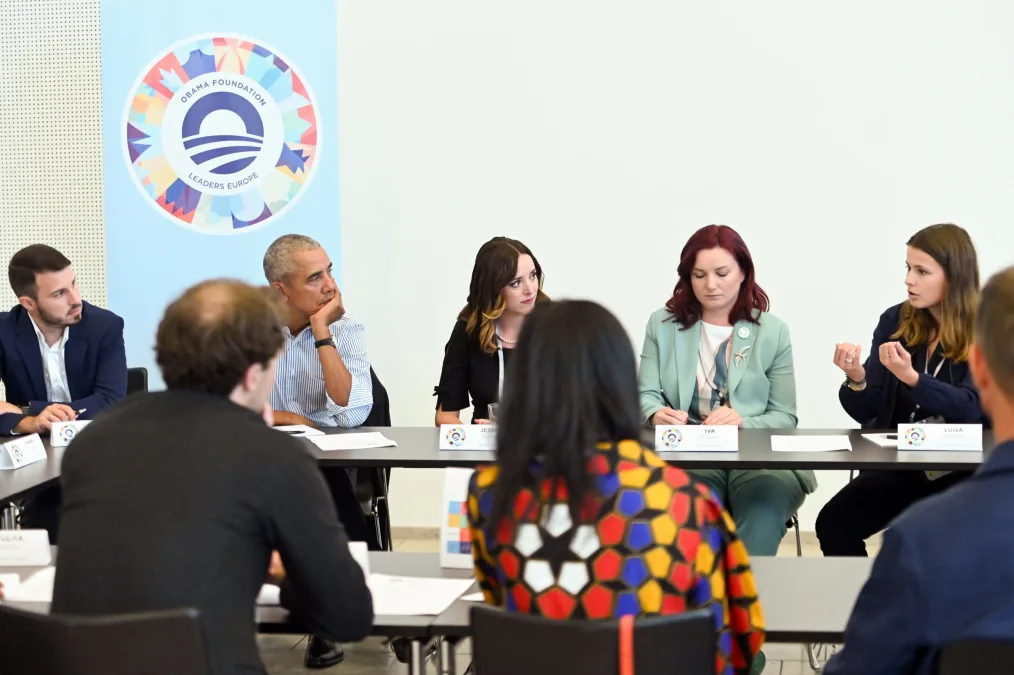 President Obama rests his face in his hand as he sits around a table with eight others. All are dressed professionally and have a range of light to medium skin tones. A colorful sign in the background reads, “Obama Foundation Leaders Europe.” A blue rising sun logo is in the center.