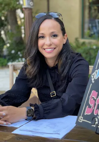 A woman with a medium-light skin tone, shoulder-length dark hair and sunglasses, is shown at a table
smiling toward the camera.