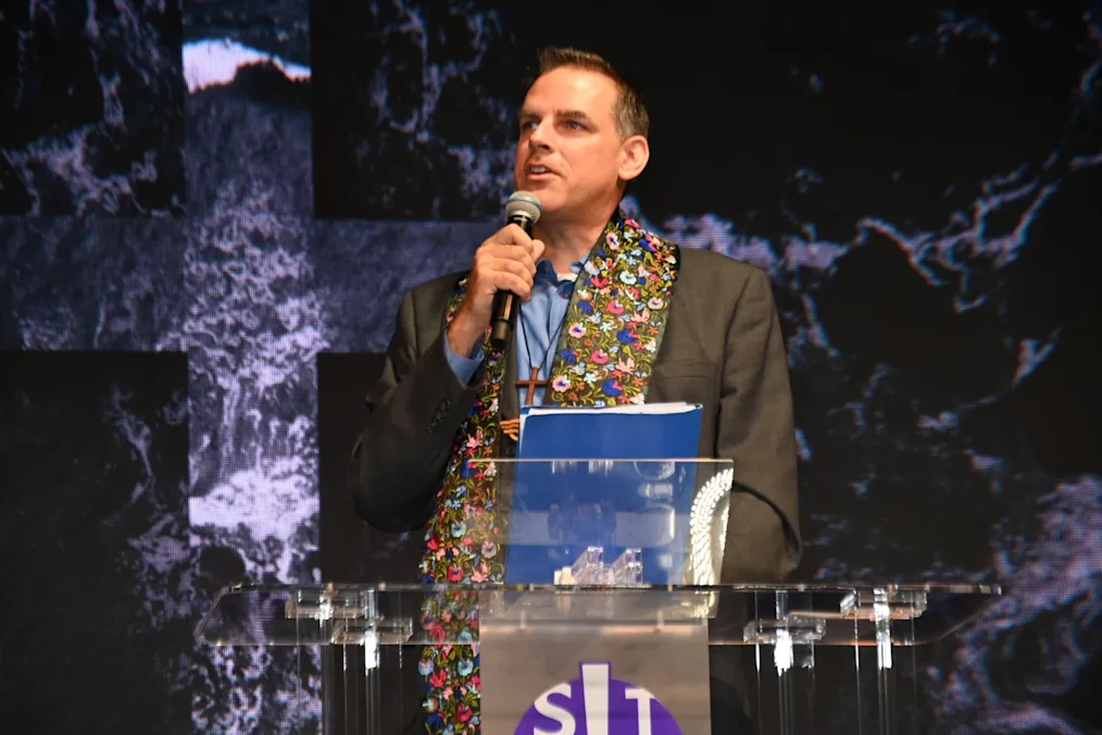 Jerad Morey, a man with a light skin tone and salt and pepper hair, speaks from a podium and holds a microphone while hosting worship service for the installation of Minnesota Council of Churches’ new CEO, Elder Suzanne P. Kelly.