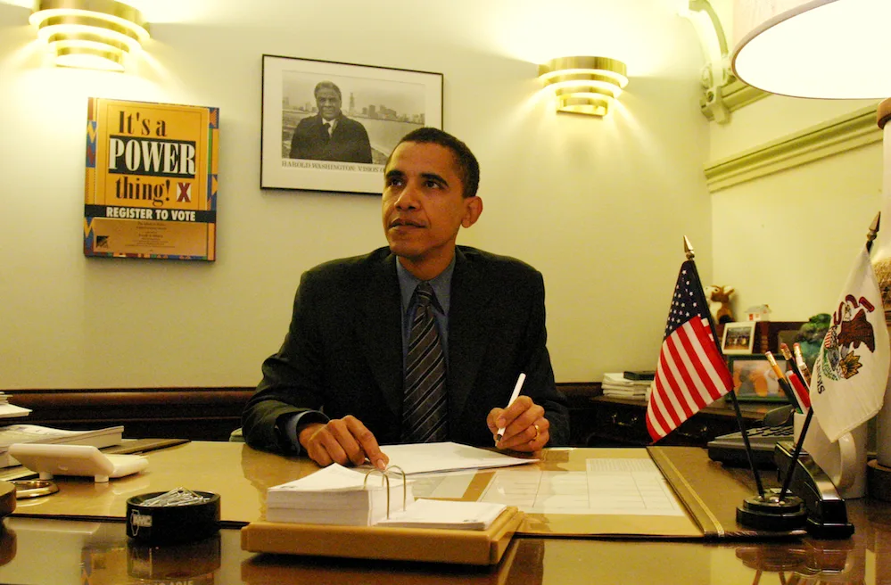 Then-Senator Barack Obama sits behind a desk looking off camera, holding a pen, with a photo of Harold Washington on the wall behind him.