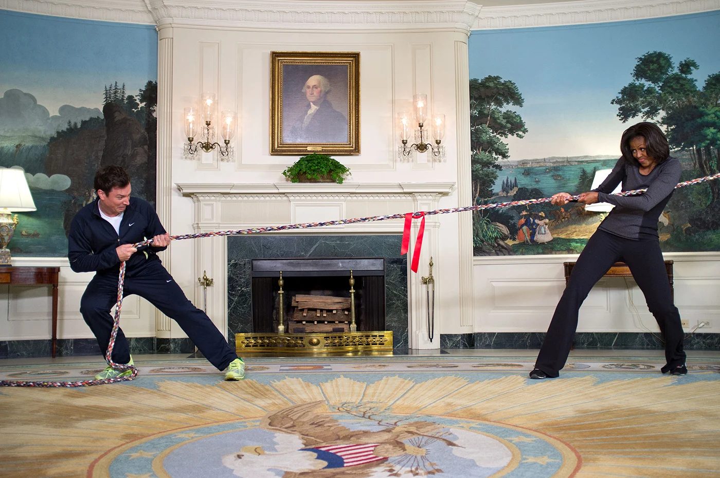 Michelle Obama plays tug-of-war with Jimmy Fallon in a large room in the White House. They are both dressed in athletic wear and a portrait of George Washington hangs above a fireplace behind them.