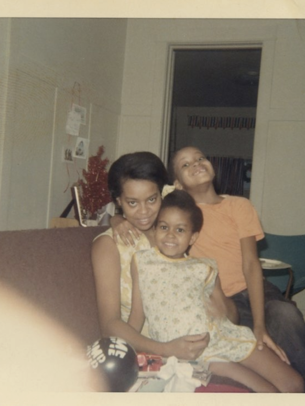 An image of Marian Robinson, Michelle Obama, and Craig Robinson sitting on a couch. The picture was photographed in the 1970s. Marian Robinson is sitting on a brown sofa and Michelle Obama is sitting in her lap. Craig Robinson is sitting on the couch beside them. They are all smiling into the camera.